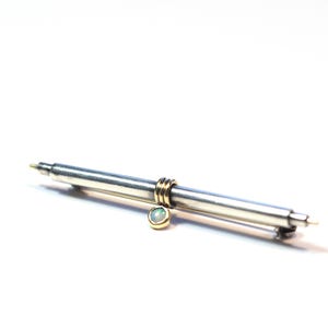 Könnte beinhalten: Eine silberne und goldene Bar-Brosche mit einem kleinen Opal-Edelstein, der in der Mitte hängt. Die Brosche besteht aus zwei Metallstäben, die mit einem goldenen Ring verbunden sind.