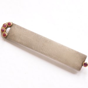 Op de afbeelding: Een zilveren en gouden staafhanger met een rij rode edelstenen aan het ene uiteinde en een enkele rode edelsteen aan het andere uiteinde.