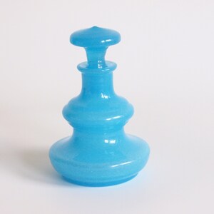 May include: A light blue glass bottle with a round, bulbous body and a round, flat stopper. The bottle has a textured surface.