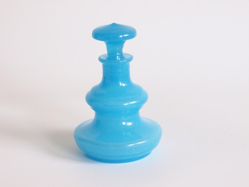 May include: A light blue glass bottle with a round, bulbous body and a round, flat stopper. The bottle has a textured surface.