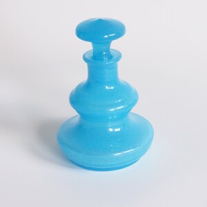 May include: A light blue glass bottle with a wide base and a rounded top. The bottle has a textured surface and a frosted finish.