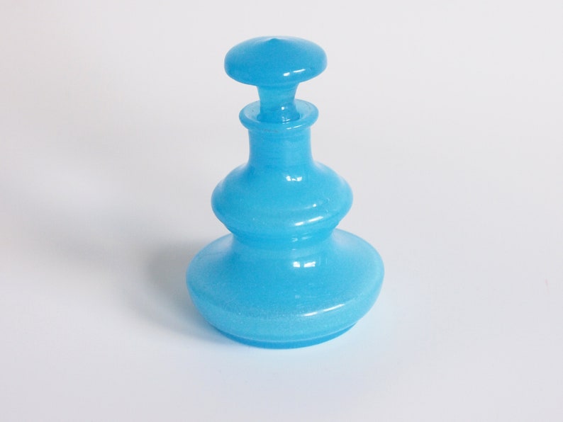 May include: A light blue glass bottle with a wide base and a rounded top. The bottle has a textured surface and a frosted finish.