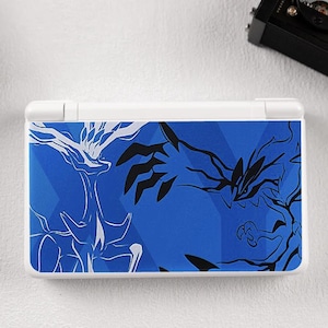 May include: A white and blue handheld gaming console featuring a blue geometric design with white and black illustrations. The console is rectangular with a hinged top and bottom. The design is reminiscent of a video game theme.