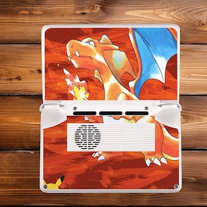 May include: White handheld gaming console featuring a vibrant orange and red graphic of a Charizard, a popular fire-type Pokémon. The console's design includes blue wings and a fiery background, with a small Pikachu figure visible.