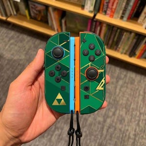 Könnte beinhalten: Ein Paar grüne Game-Controller mit goldenem geometrischem Muster und goldenem Triforce-Emblem. Der linke Controller hat eine hellblaue Seitenschiene, der rechte eine orangefarbene, beide mit befestigten schwarzen Handgelenkschlaufen.