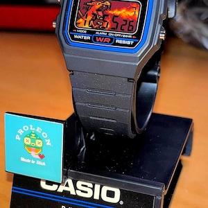 May include: A black Casio F-91W digital watch with a custom pixel art display showing a fiery scene. The watch is mounted on a black display stand featuring the Casio logo, product specifications, and a small square sticker with a pixelated character.