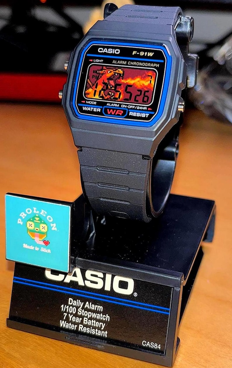 May include: A black Casio F-91W digital watch with a custom pixel art display showing a fiery scene. The watch is mounted on a black display stand featuring the Casio logo, product specifications, and a small square sticker with a pixelated character.