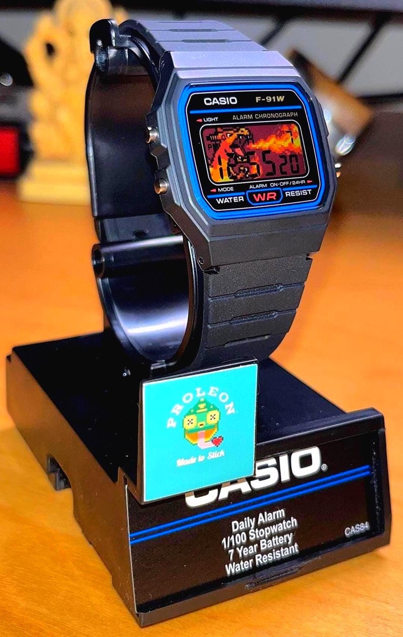 May include: A black Casio F-91W digital watch with a blue bezel, featuring a digital display showing a graphic of a fire-breathing creature. The watch is mounted on a black display stand with the text Daily Alarm, 1/100 Stopwatch, 7 Year Battery.