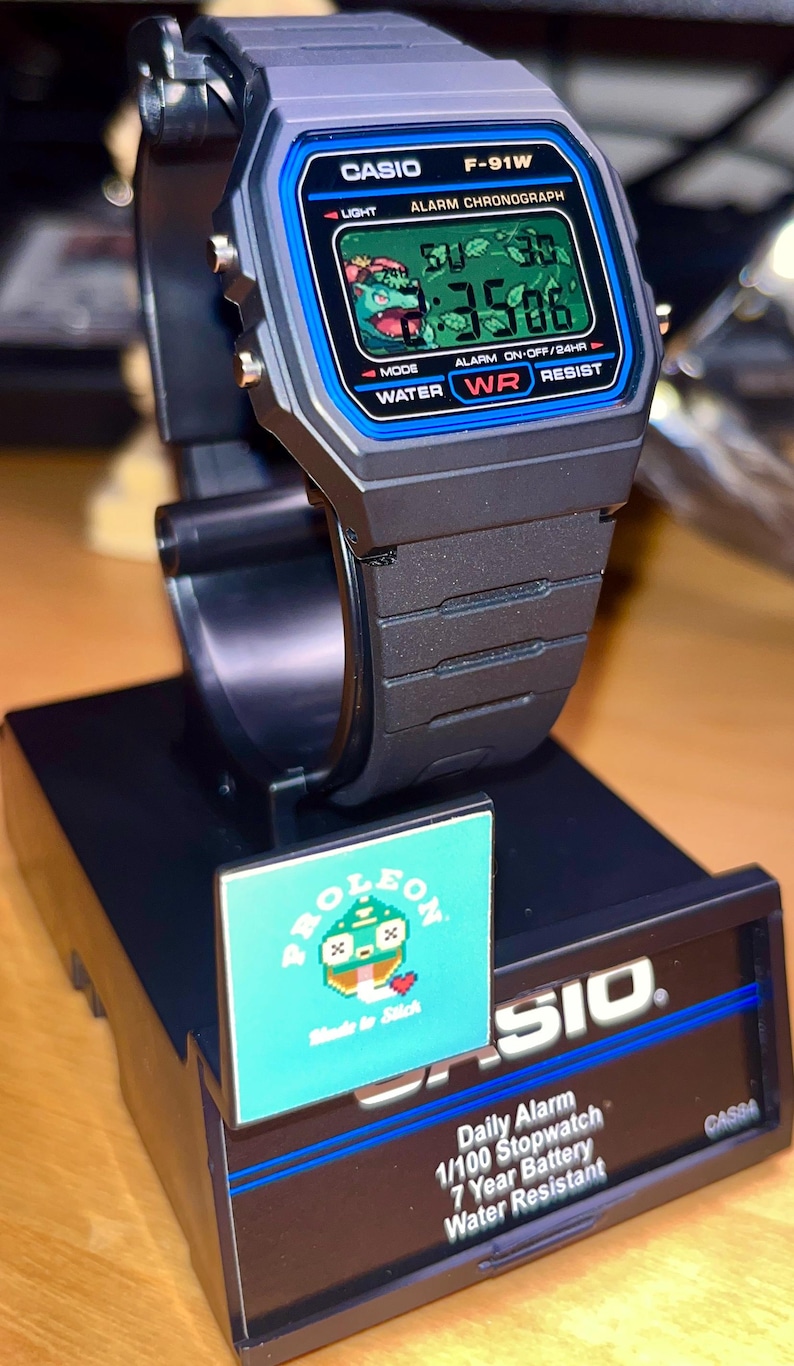 May include: A black Casio F-91W digital watch with a blue bezel and a custom graphic of a green creature on the LCD screen. The watch is mounted on a black display stand featuring the text CASIO, Daily Alarm, 1/100 Stopwatch, 7 Year Battery, and Water Resistant.