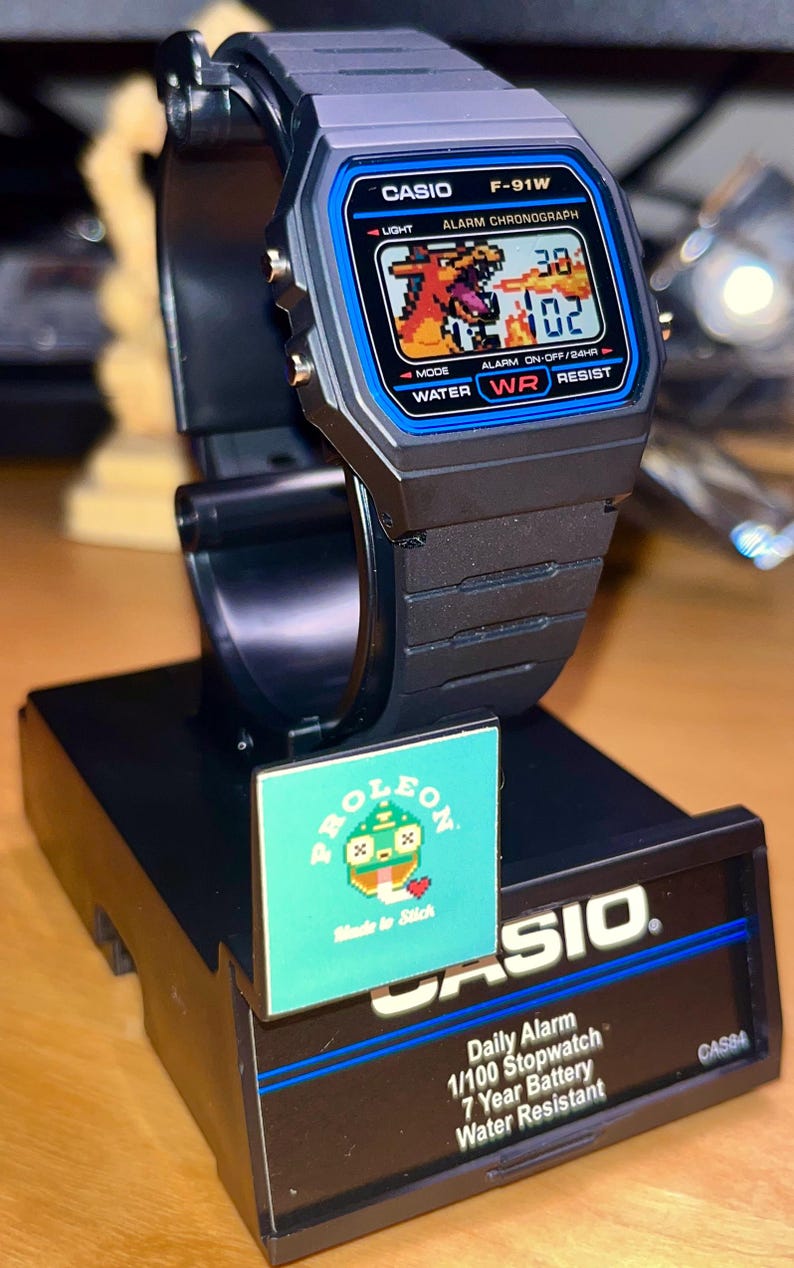 May include: A black Casio F-91W digital watch with a blue bezel and a pixelated Charizard graphic on the LCD screen. The watch is mounted on a black display stand featuring the text CASIO, Daily Alarm, 1/100 Stopwatch, 7 Year Battery, Water Resistant.
