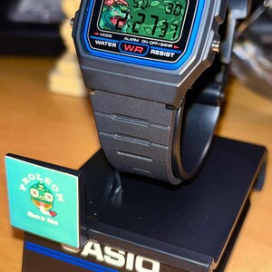 May include: A black Casio F-91W digital watch with a blue border around the screen, featuring a pixelated green creature graphic on the display. The watch is mounted on a black display stand with the text CASIO, Daily Alarm, 1/100 Stopwatch, 7 Year Battery, Water Resistant, and CAS84.