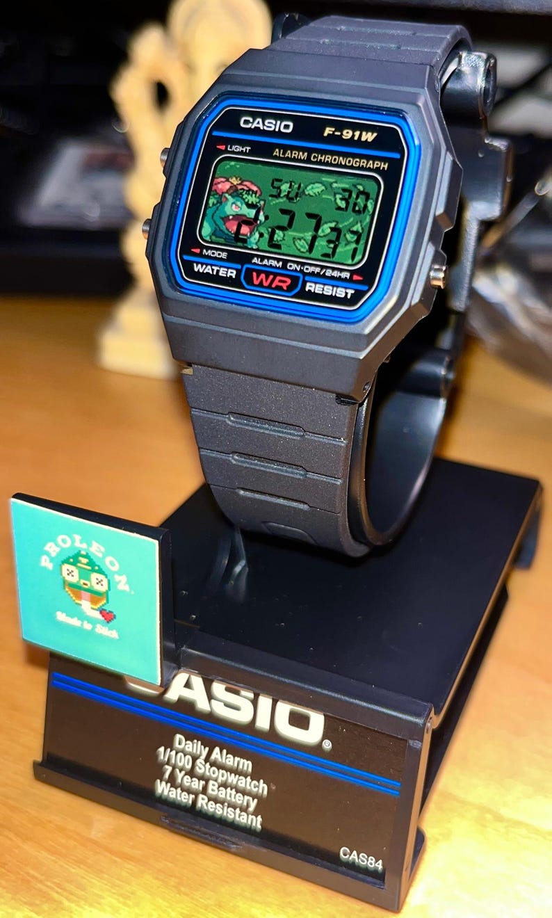 May include: A black Casio F-91W digital watch with a blue border around the screen, featuring a pixelated green creature graphic on the display. The watch is mounted on a black display stand with the text CASIO, Daily Alarm, 1/100 Stopwatch, 7 Year Battery, Water Resistant, and CAS84.