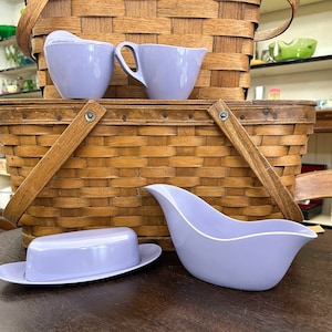 May include: A set of four lavender-colored ceramic serving dishes. The dishes include a creamer, a sugar bowl, a butter dish, and a gravy boat. The dishes are arranged on top of a wicker picnic basket.