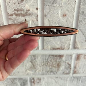 May include: A copper-colored cabinet pull with an ornate design. The pull is oval-shaped and has a black background with a floral pattern.