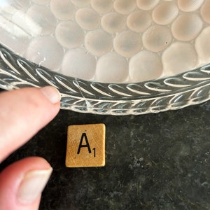 May include: A close-up of a clear glass bowl with a textured, bubble-like pattern. A wooden Scrabble tile with the letter "A" and the number "1" is next to the bowl. The bowl's rim has a leaf design.