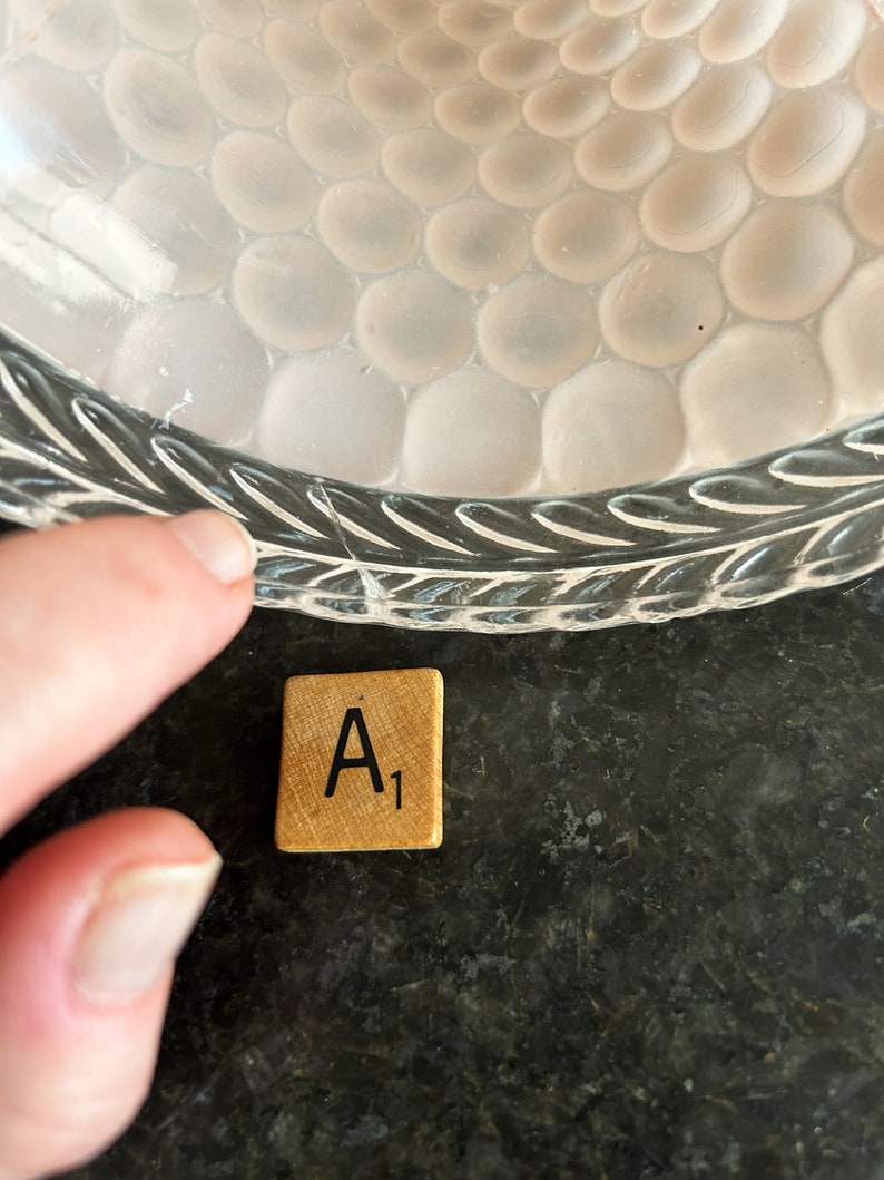 May include: A close-up of a clear glass bowl with a textured, bubble-like pattern. A wooden Scrabble tile with the letter "A" and the number "1" is next to the bowl. The bowl's rim has a leaf design.
