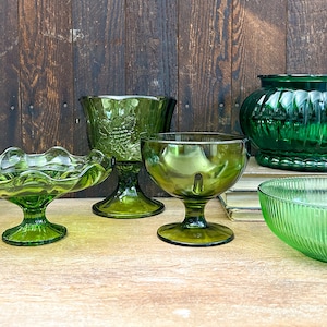 May include: A collection of six green glass vessels including a textured bowl, a ruffled pedestal dish, a footed goblet with grape motifs, a smooth footed bowl, a large ribbed bowl, and a wide, rounded bowl with vertical fluted detailing.