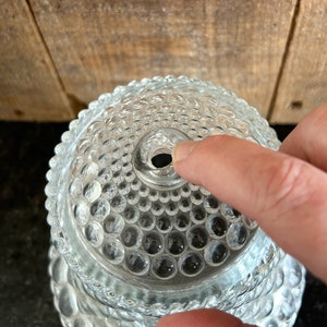 May include: Clear glass hobnail-patterned lid with a central hole. The lid has a rounded shape and is covered in small, raised, circular bumps. The lid is partially obscuring the background, which appears to be a wooden surface.