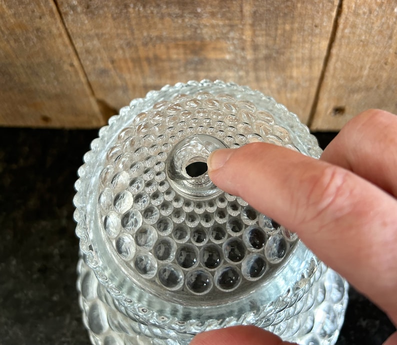 May include: Clear glass hobnail-patterned lid with a central hole. The lid has a rounded shape and is covered in small, raised, circular bumps. The lid is partially obscuring the background, which appears to be a wooden surface.