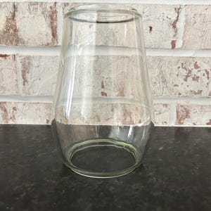 May include: A clear, glass, bell-shaped vase with a wide base and a narrow top. The vase is empty and sits on a dark surface, with a white brick wall in the background.