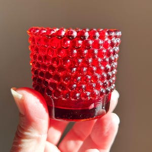 May include: A small, ruby red glass votive candle holder. The exterior is textured with a pattern of small, raised bumps. The candle holder is held in a person's hand, and the light catches the glass, creating highlights and shadows.