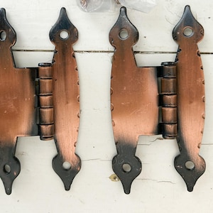 May include: Two antique copper-colored hinges with black accents. Each hinge has a decorative shape with a pointed top and bottom, and a series of rivets along the sides. The hinges are designed for a door or cabinet.