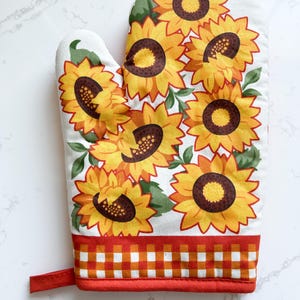May include: White oven mitt featuring a vibrant sunflower pattern. The mitt has a red and orange gingham border and a red loop for hanging. The sunflowers are yellow and orange with brown centers and green leaves.