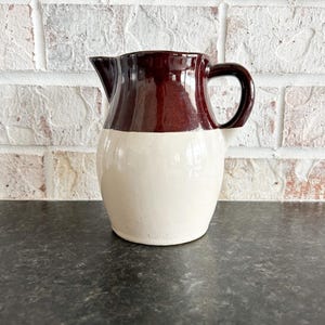 May include: A brown and white glazed ceramic pitcher with a handle. The pitcher has a rounded body and a wide mouth.