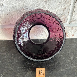 May include: A deep purple glass candle holder with a circular design. The holder features intricate embossed patterns and a clear center. A wooden Scrabble tile with the letter "B" and the number "3" is in the foreground.