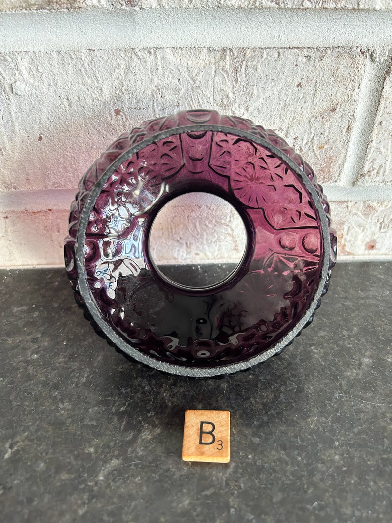 May include: A deep purple glass candle holder with a circular design. The holder features intricate embossed patterns and a clear center. A wooden Scrabble tile with the letter "B" and the number "3" is in the foreground.