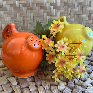 May include: Two ceramic pig salt and pepper shakers. The salt shaker is orange and the pepper shaker is yellow. They are sitting on a woven wicker surface.