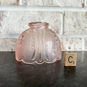 May include: A pink glass lampshade with a floral and diamond pattern. The lampshade is dome-shaped and sits on a dark surface. A wooden tile with the letter "C" and the number "3" is next to the lampshade.