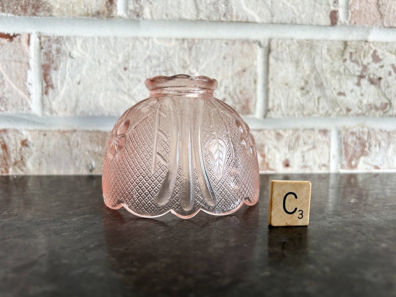 May include: A pink glass lampshade with a floral and diamond pattern. The lampshade is dome-shaped and sits on a dark surface. A wooden tile with the letter "C" and the number "3" is next to the lampshade.