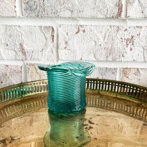 May include: A teal blue glass candle holder shaped like a hat. The holder has a spiral design and is sitting on a gold tray.