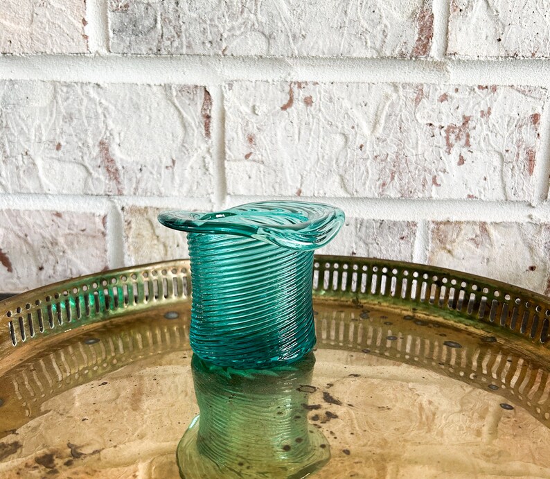 May include: A teal blue glass candle holder shaped like a hat. The holder has a spiral design and is sitting on a gold tray.