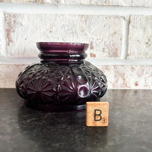 May include: A deep purple, vintage-style glass inkwell with a detailed, textured pattern. The inkwell has a round base and a flared top. A wooden tile with the letter "B" and the number "3" is next to the inkwell.