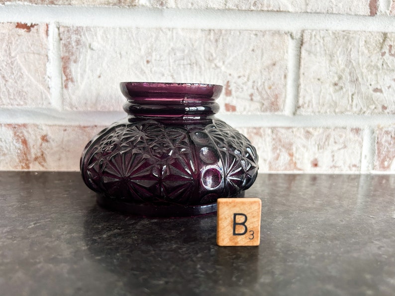 May include: A deep purple, vintage-style glass inkwell with a detailed, textured pattern. The inkwell has a round base and a flared top. A wooden tile with the letter "B" and the number "3" is next to the inkwell.