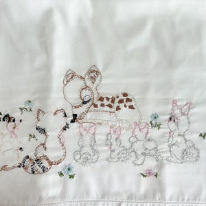 May include: White fabric with embroidered woodland animals, including a deer with brown spots, raccoons, and rabbits. The embroidery features pink, blue, and brown thread, creating a whimsical design. The fabric has a subtle sheen.