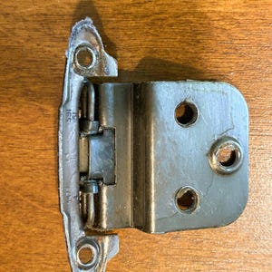 May include: A silver-toned metal cabinet hinge featuring a wrap-around design with a central pivot point. The hinge has three circular mounting holes on the flat plate and two additional holes on the curved flange, with the text 591 51 visible.