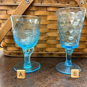 May include: Two vintage, light blue goblets. One goblet has a leaf design, the other has a diamond pattern. Both have a stem and a round base. The goblets are on a dark brown surface, with a wicker basket in the background.