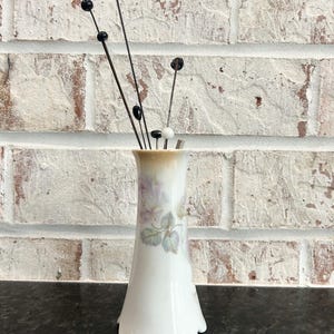 Puede incluir: Un jarr&oacute;n de porcelana blanca con base festoneada, adornado con un dise&ntilde;o floral en tonos morados y verdes. El jarr&oacute;n contiene varios tallos decorativos con cuentas negras y blancas, creando una exhibici&oacute;n delicada y elegante.