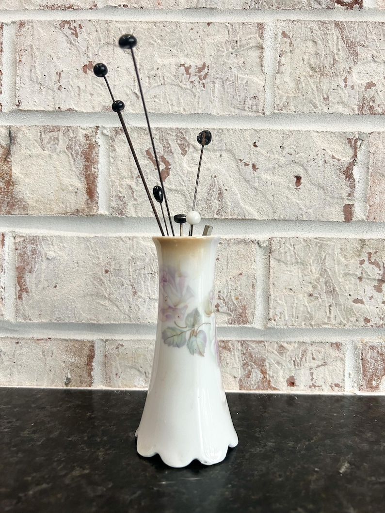 Puede incluir: Un jarr&oacute;n de porcelana blanca con base festoneada, adornado con un dise&ntilde;o floral en tonos morados y verdes. El jarr&oacute;n contiene varios tallos decorativos con cuentas negras y blancas, creando una exhibici&oacute;n delicada y elegante.