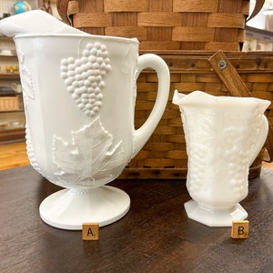 May include: Two white milk glass pitchers with a grapevine design. The larger pitcher has a handle and a base. The smaller pitcher has a handle and a base. Both pitchers are labeled with letter tiles, A1 and B3.