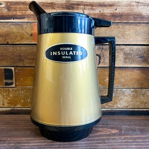 May include: A gold-colored double-walled insulated pitcher with a black lid and handle. The pitcher has a black oval label that reads "DOUBLE INSULATED WALL".