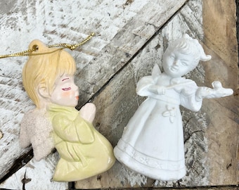 May include: Two angel ornaments. One is light yellow with closed eyes and praying hands. The other is white and playing a violin. Both have wings and are on a rustic wooden surface. Gold string hangers are attached.