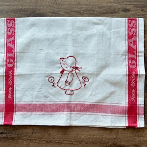 May include: White linen kitchen towel with red accents and embroidery. Features a red border and vertical stripes with the word "GLASS" in red. The center has a red embroidered girl and flowers.