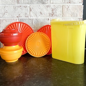 May include: A set of four vintage orange and red plastic bowls and a yellow plastic pitcher. The bowls are stacked on top of each other and the pitcher is standing upright.