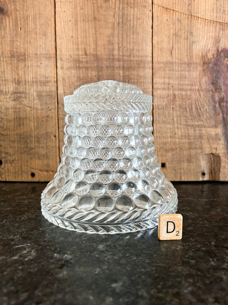 May include: Clear glass jar with a textured, bubble-like pattern. The jar has a lid with a decorative rope-like design around the top. The base of the jar has a similar design. A wooden tile with the letter "D" and the number "2" is next to the jar.
