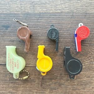 May include: A collection of six plastic whistles in various colors, including brown, black, yellow, and red. Some whistles have text, and one has a chain. The whistles are arranged on a wooden surface.