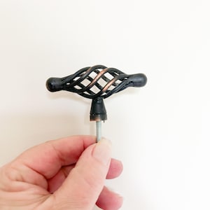 May include: A T-shaped cabinet knob featuring a matte black finish with exposed copper-toned metal accents within the twisted, cage-like center design. The hardware includes a threaded metal bolt extending from the base for installation purposes.